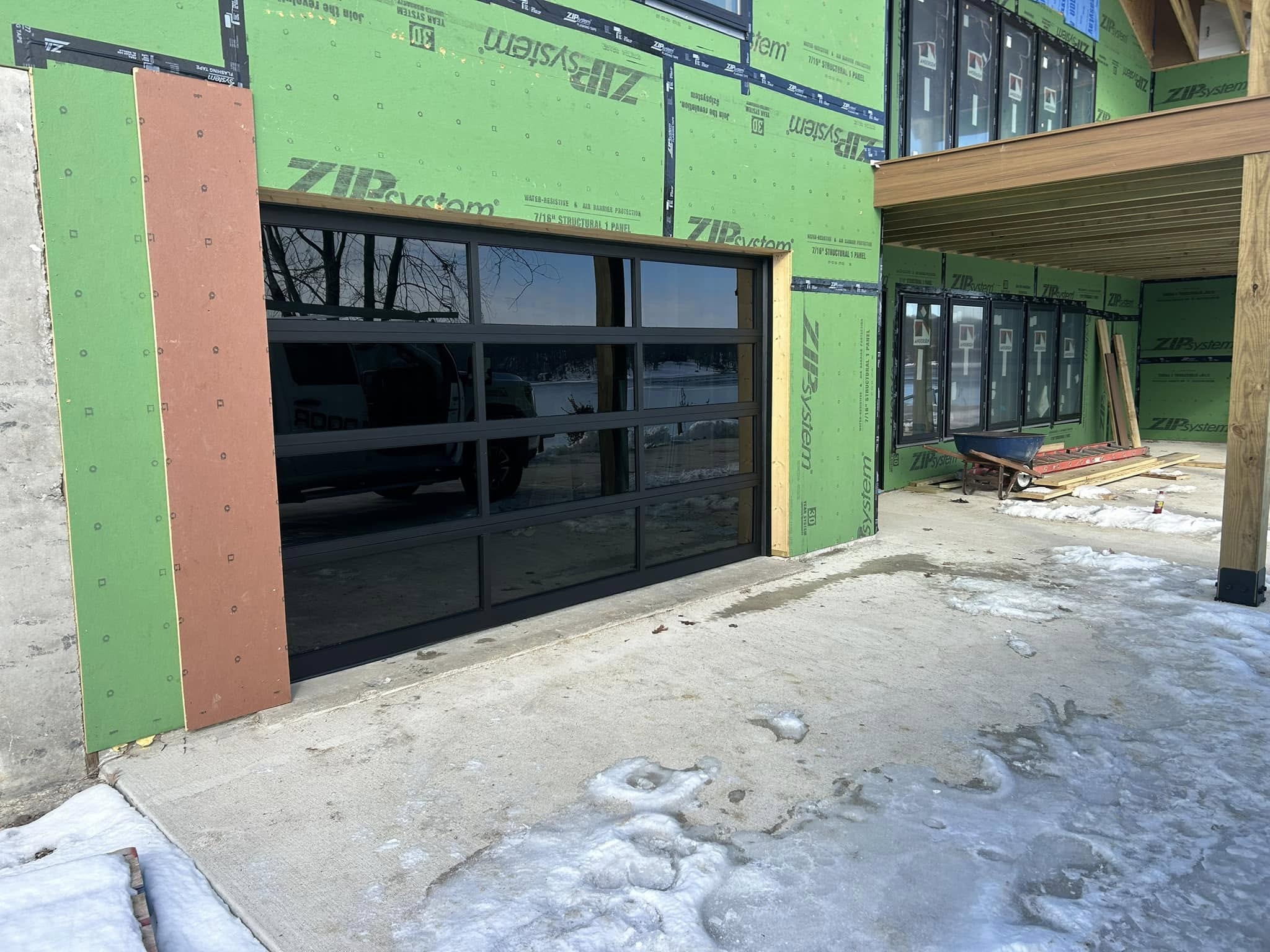 Glass-panel garage door on a house under construction with green ZIP System sheathing and snow on the ground.