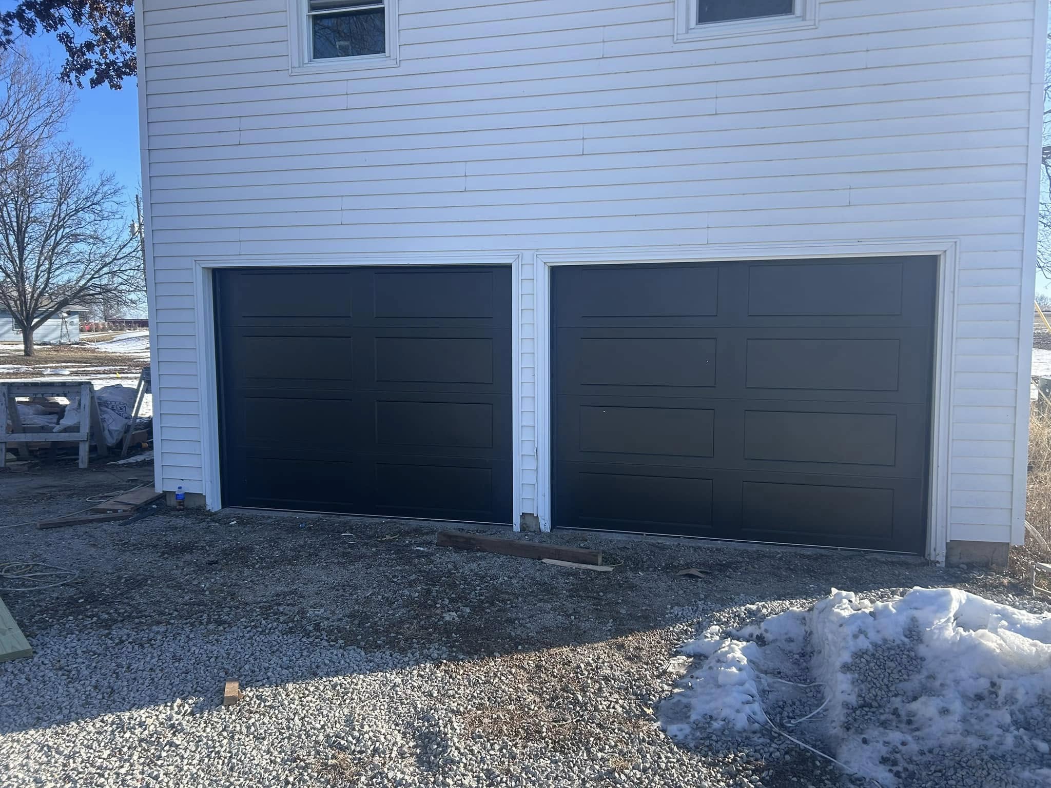 Two dark garage doors on a white house with gravel driveway and patches of snow.